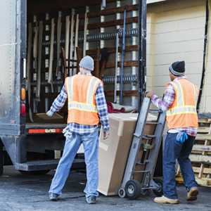 Two male workers in their 40s at the back of a truck, loading or unloading a large cardboard box. The men are wearing plaid shirts, reflective vests and jeans.